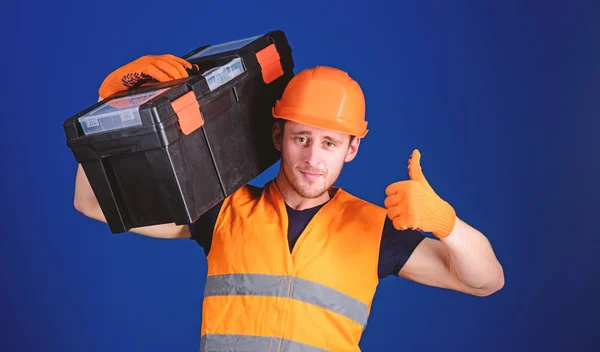 Homem de capacete, chapéu duro segura caixa de ferramentas e mostra gesto polegar para cima, fundo azul. Trabalhador, reparador, reparador, construtor no rosto confiante carrega caixa de ferramentas no ombro. Conceito de consulta de reparação — Fotografia de Stock