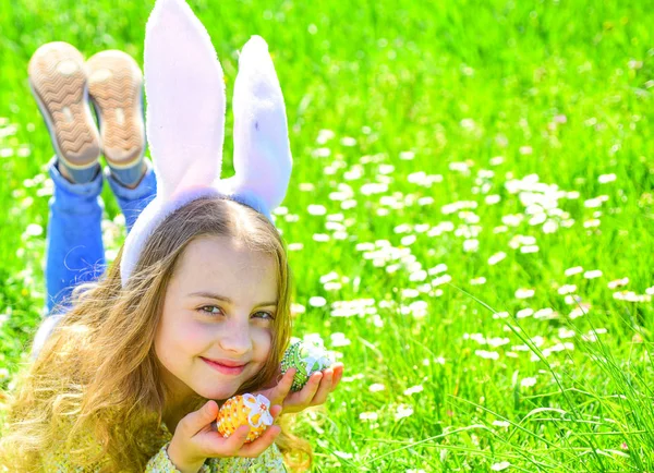 Child with cute bunny ears lying at meadow. Little girl hunting for Easter egg in spring garden on Easter day, traditional celebration. Easter feast concept. Cute child lay on grass with egg in hands