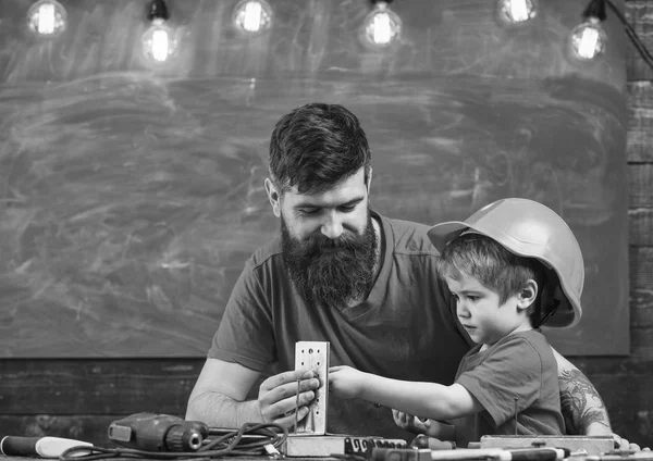 髭のツールを使用して、背景に黒板の教室で少し息子を教える父。少年は、保護用のヘルメットで忙しい子になります、手でお父さんと修復します。父親のアシスタントのコンセプト — ストック写真