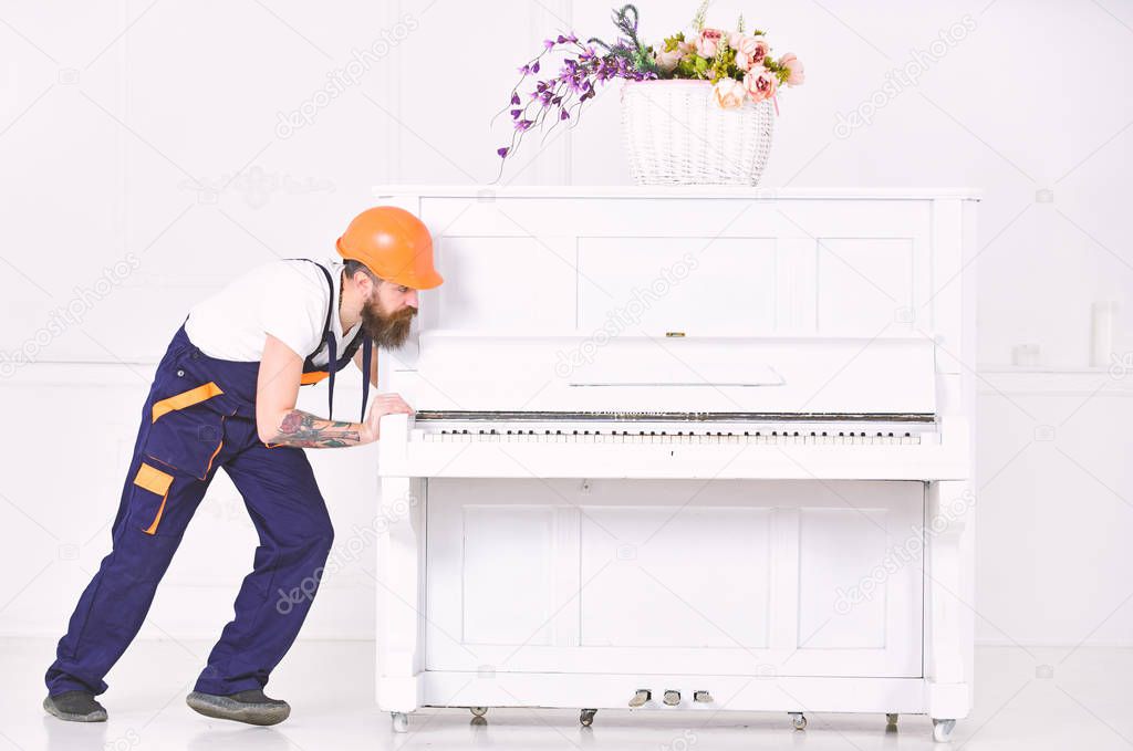 Handsome bearded strong man moving piano with open keyboard and glass vase with flowers on white background. Home service concept
