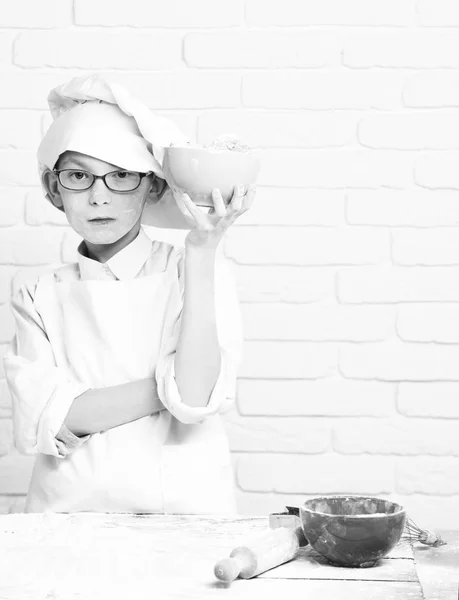 Jovem menino bonito cozinheiro chef em uniforme branco e chapéu na farinha de rosto manchado com óculos de pé perto da mesa com rolo de pino e segurando tigela turquesa com biscoitos de chocolate no fundo da parede de tijolo — Fotografia de Stock