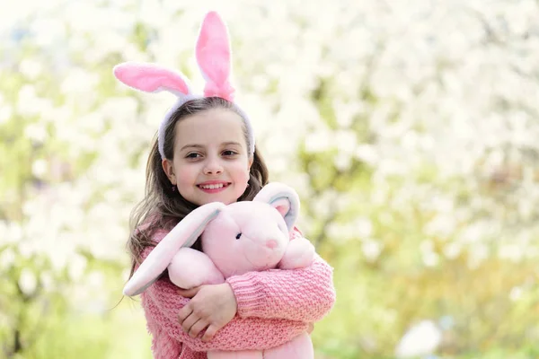 Påsk och våren. Glad tjej håller rosa kanin i trädgården med blommande träd. Barndom, ungdom och tillväxt. Barnet ler med bunny öron pannband på långt hår. Semester firande koncept — Stockfoto