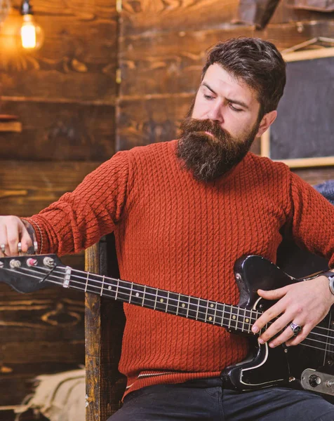 Músico de rock com aparência brutal posando com instrumento. Homem barbudo afinando guitarra elétrica. Homem com barba elegante e bigode em camisola de malha terracota e calça jeans preta relaxante, conceito de música — Fotografia de Stock