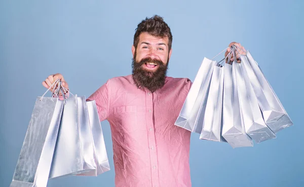 Hombre de compras en temporada de ventas con descuentos. Concepto de venta y descuento. El hombre con barba y bigote sostiene bolsas de compras, fondo azul claro. Hipster en la cara feliz es adicto a las compras o adicto a las compras — Foto de Stock