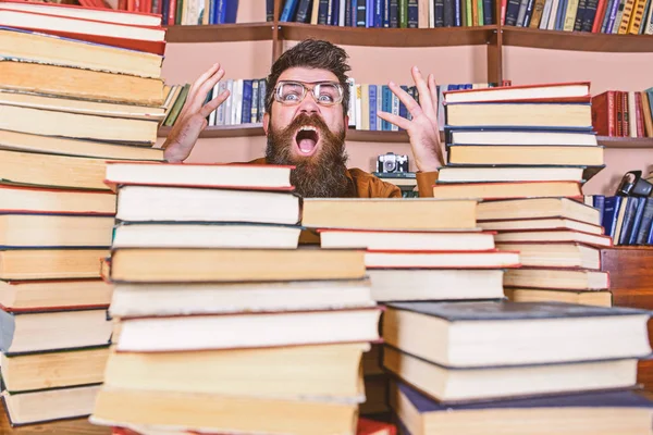 Mann mit schockiertem Gesicht zwischen Bücherstapeln in der Bibliothek, Bücherregale im Hintergrund. Lehrer oder Schüler mit Bart tragen Brillen, sitzen mit Büchern am Tisch und sind defokussiert. wissenschaftliches Entdeckungskonzept — Stockfoto