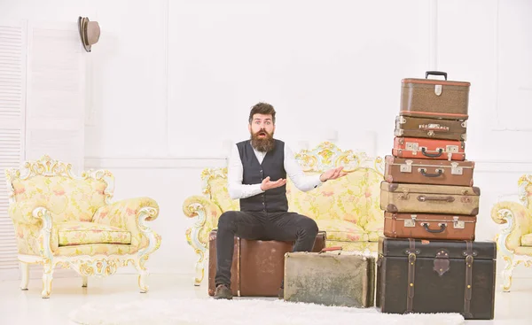 Macho-Eleganz auf überraschtem Gesicht sitzt verwirrt am Ende des Packens neben einem Stapel alter Koffer. Gepäck- und Umzugskonzept. Mann mit Bart und Schnurrbart gepacktes Gepäck, weißer Innenhintergrund — Stockfoto
