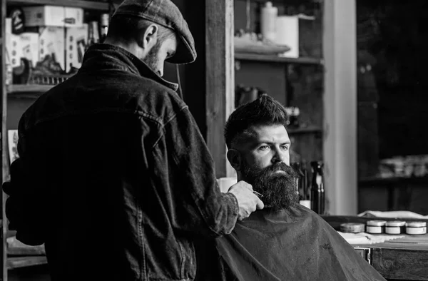 Hipster avec barbe recouverte d'une cape servant par un coiffeur professionnel dans un élégant salon de coiffure. Concept de toilettage. Barbier occupé avec la barbe de toilettage du client hipster, fournitures de beauté sur fond — Photo