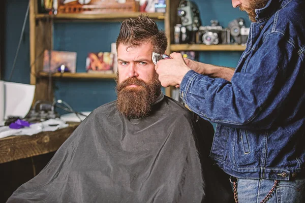 Barber with clipper trimming hair on temple of client. Hipster lifestyle concept. Barber with hair clipper works on hairstyle for bearded man barbershop background. Hipster client getting haircut
