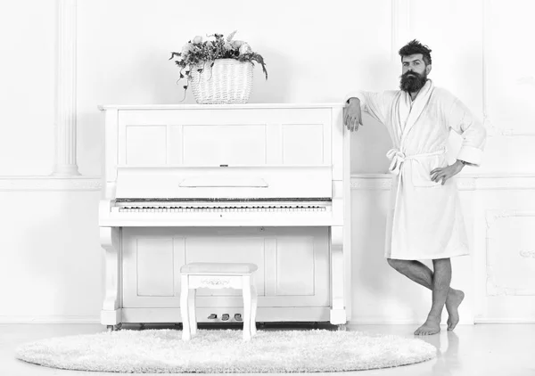 El hombre con barba en albornoz disfruta de la mañana mientras está de pie cerca del piano. Concepto de músico con talento. Hombre soportes serios y se apoya en el instrumento musical de piano en el interior blanco sobre fondo —  Fotos de Stock