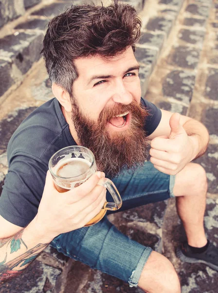 Homem com barba e bigode segura vidro com cerveja e mostra polegar para cima, fundo escadas de pedra. Hipster na cara alegre beber cerveja de esboço ao ar livre. Macho descansando com cerveja. Sexta-feira relaxar conceito — Fotografia de Stock