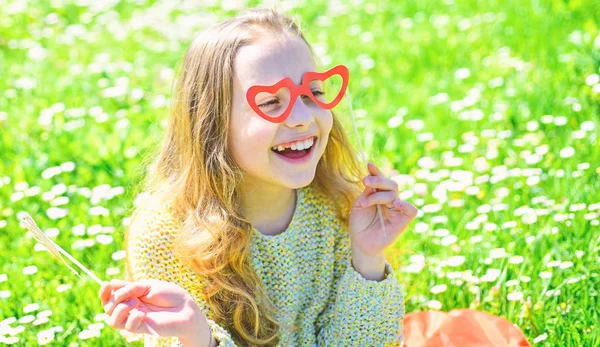 Love from first sight concept. Child posing with cardboard heart shaped eyeglasses. Girl on happy face spend leisure outdoors. Girl sits on grass at grassplot, green background