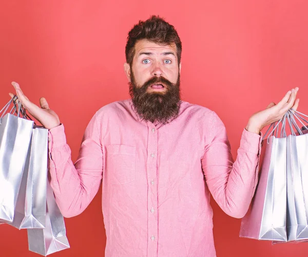 Male shopaholic with long beard isolated on red background. Hipster in pink shirt with stylish beard holding silver shopping bags. Bearded man with surprised look spent all his money on sale