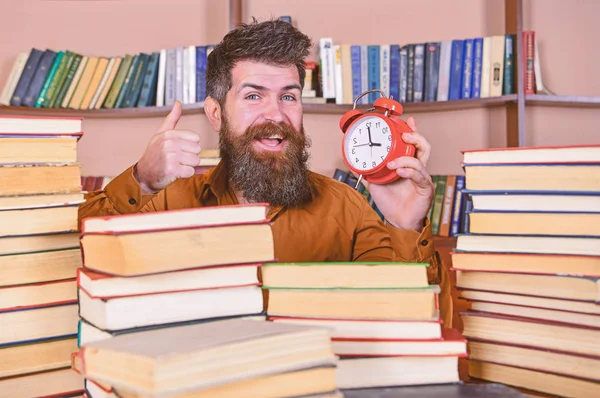 Man, vetenskapsman som kikar ut ur högar av böcker med väckarklocka. Lärare med skägg studera i biblioteket. Tid flow-konceptet. Mannen på glada ansikte innehar klocka, visar tummen upp, bokhyllor på bakgrund — Stockfoto