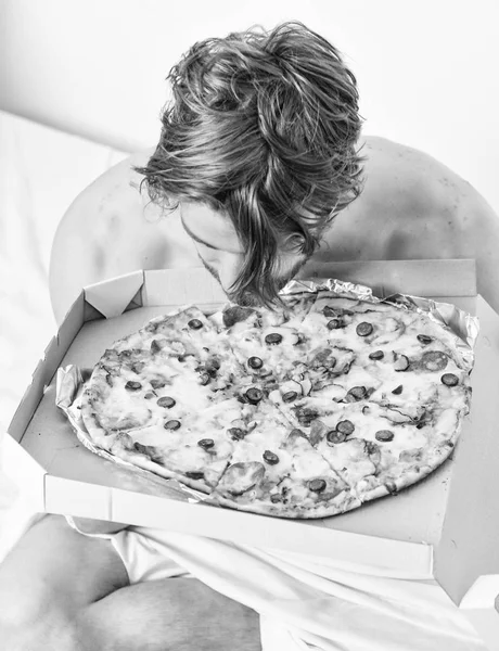 Homme barbu beau gars manger de la nourriture au fromage pour le petit déjeuner au lit. Jeune homme torse nu avec pizza au lit. Étudiant est à la maison sur le lit dans un appartement lumineux manger une délicieuse pizza . — Photo