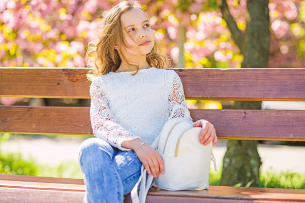 Girl on dreamy face sits on bench, sakura tree on background, defocused. Spring walks concept. Cute child with backpack enjoy sunny spring day. Girl relaxing while walk in park near cherry blossom