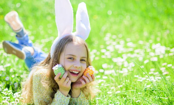 Chica en la cara feliz sostiene huevos de Pascua en las manos, hierba en el fondo. Niño con orejas de conejo acostado en el prado, espacio de copia. Chica con el pelo largo acostado en la parcela de hierba en el soleado día de primavera. Concepto Pascua — Foto de Stock