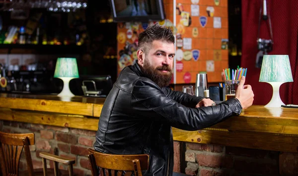 Brutal hipster bearded man sit at bar counter drink beer. Friday evening. Hipster relaxing at bar with beer. Bar is relaxing place to have drink and relax. Man with beard spend leisure in dark bar