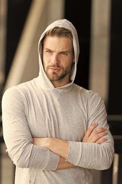 Cool and confident. Man unshaven hooded folded arms looks handsome and cool. Guy bearded attractive cares about appearance. Man bristle serious face, urban background, defocused. Metrosexual concept