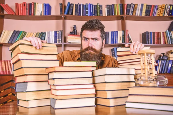 L'enseignant ou l'élève avec la barbe est assis à table avec des livres, déconcentré. Homme au visage sérieux entre des piles de livres, tout en étudiant à la bibliothèque, étagères de bibliothèque sur fond. Concept bibliophile — Photo