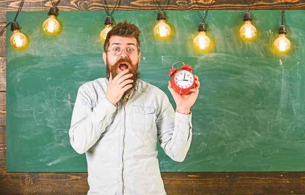 O hipster barbudo segura relógio, quadro no fundo, espaço de cópia. Esqueci-me do conceito do tempo. O professor de óculos tem despertador. Homem com barba e bigode no rosto chocado na sala de aula — Fotografia de Stock