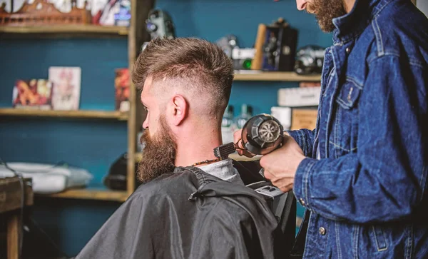 Barbershop concept. Hipster bearded client got hairstyle. Barber with hairdryer blows off hair out of cape. Barber with hairdryer works on hairstyle for bearded man, barbershop background — Stock Photo, Image