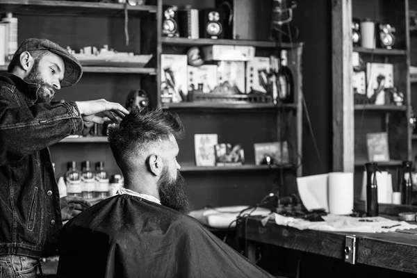 Barber styling hair of bearded client with wax. Hipster client getting hairstyle. Barbershop concept. Man with beard and mustache sits in hairdressers chair in front of mirror, barbershop background — Stock Photo, Image