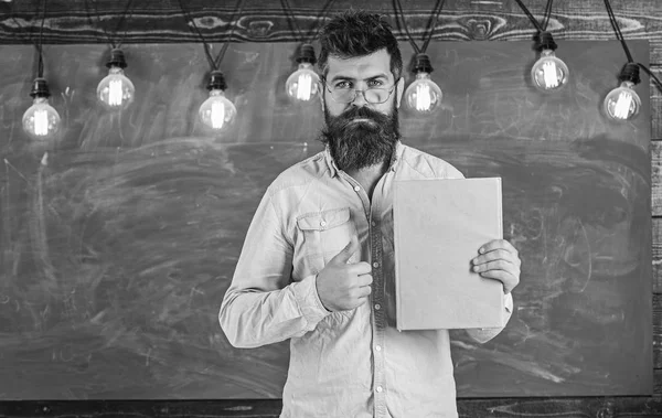 Bearded hipster holds book, chalkboard on background. Teacher in eyeglasses recommends book, thumbs up. Recommendation concept. Man with beard and mustache on serious face stand in front of chalkboar — Stock Photo, Image