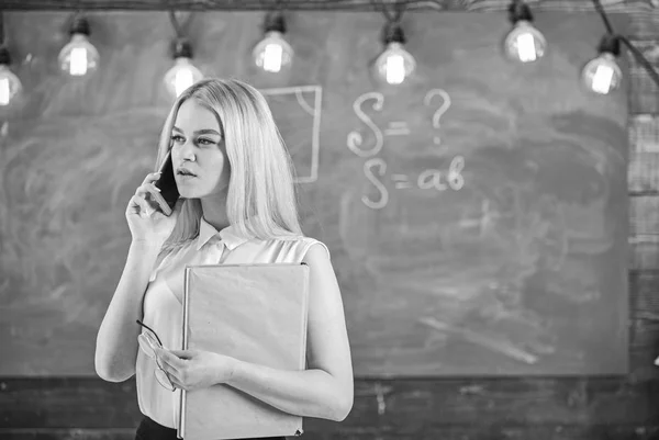 Mujer con libro hablando por teléfono móvil en clase. Profesor reportando sobre estudiantes ausentes. Profesor en problemas concentrados de resolución de rostros en el teléfono, pizarra en el fondo. Concepto de informe móvil — Foto de Stock