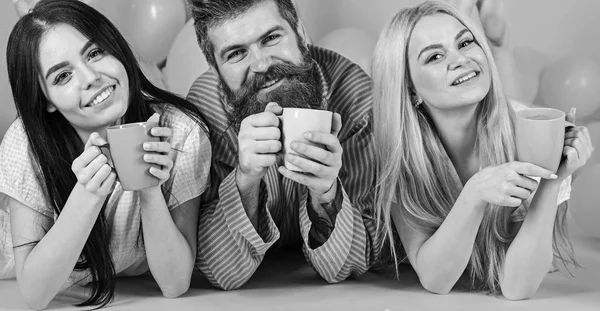 Hombres y mujeres con ropa de hogar, pijamas. Trío relajarse en la mañana con café. Amantes de tomar café en la cama. Amantes en concepto de cama. Hombre y mujeres, amigos en caras felices, fondo rosa — Foto de Stock