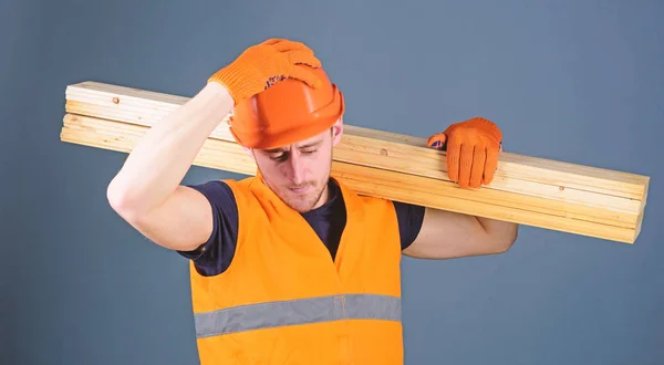 Concepto de seguridad y protección. Hombre con casco, sombrero duro y guantes de protección sostiene la viga de madera, fondo gris. Carpintero, carpintero, fuerte constructor en la cara seria lleva viga de madera en el hombro —  Fotos de Stock