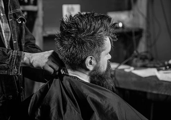 Man with beard and mustache sits in barbershop, beauty supplies on background. Hipster with beard waits while barber cover neck with cape. Barbershop concept. Man bearded client of hipster barbershop — Stock Photo, Image