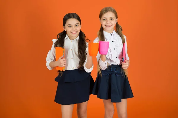 Des camarades de classe se relaxent avec un verre. Profite d'être élève. Enfants filles uniforme scolaire fond orange. Écolière tenir livre ou bloc-notes et tasse. La routine scolaire. Détends-toi. Boire du thé pendant la pause — Photo