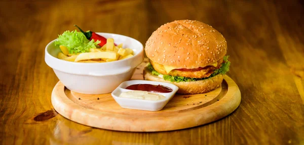 Comida de engaño. Deliciosa hamburguesa con semillas de sésamo. Hamburguesa con carne de queso y ensalada. Concepto de comida rápida. Menú de hamburguesas. Snack alto en calorías. Hamburguesa y papas fritas y salsa de tomate sobre tabla de madera —  Fotos de Stock