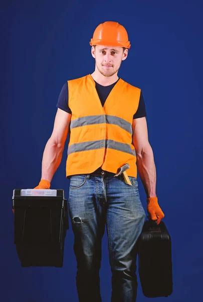 Handyman, reparador no rosto estrito vai e carrega sacos com equipamento profissional. Conceito de reparador profissional. Homem de capacete, chapéu duro segura caixa de ferramentas e mala com ferramentas, fundo azul — Fotografia de Stock