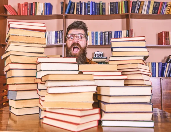 Nerd-Konzept. Lehrer oder Schüler mit Bart tragen Brillen, sitzen mit Büchern am Tisch und sind defokussiert. Mann, Nerd auf überraschtem Gesicht zwischen Bücherstapeln in Bibliothek, Bücherregale im Hintergrund — Stockfoto