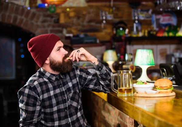 Man with beard drink beer eat burger menu. Enjoy meal in pub. High calorie snack. Brutal hipster bearded man sit at bar counter. Hipster relaxing at pub. Pub is relaxing place to have drink and relax