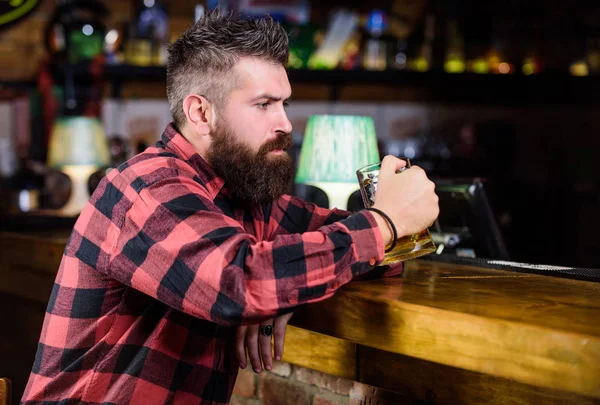 Man with beard spend leisure in dark bar. Hipster relaxing at bar with beer. Brutal hipster bearded man sit at bar counter drink beer. Order alcohol drink. Bar is relaxing place have drink and relax