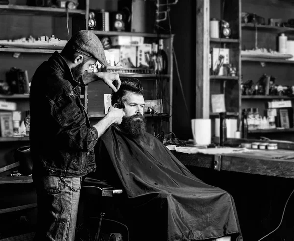 Hipster client getting haircut. Haircut concept. Barber with hair clipper works on hairstyle for man with beard, barbershop background. Barber styling hair of bearded client with comb and clipper