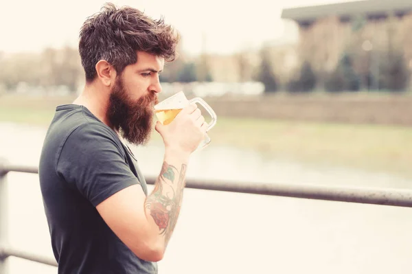 Bearded man holds beer mug, drinks beer outdoor. Man with long beard looks relaxed. Man with beard and mustache on calm face, river background, defocused. Craft beer concept