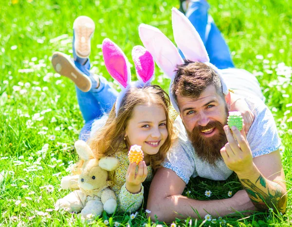 Koncepce rodinné dovolené. Otec a dcera na šťastné tváře pořádá velikonoční vajíčka, tráva na pozadí. Dívka s tátou ležící na trávě na slunečný den. Dítěte a muž s roztomilý zajíček uši hrát spolu — Stock fotografie