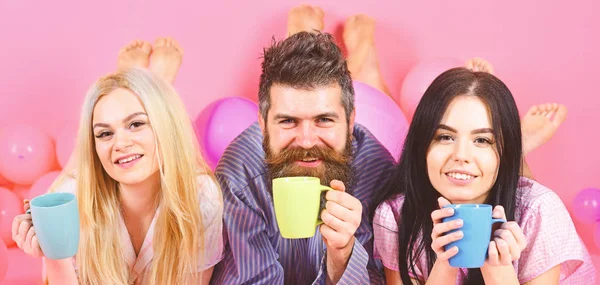 Trío relajarse en la mañana con café. Concepto de amantes. Hombres y mujeres, amigos en rostros sonrientes, fondo rosa. Hombres y mujeres con ropa de hogar, pijamas. Los amantes del amor beben café en la cama — Foto de Stock