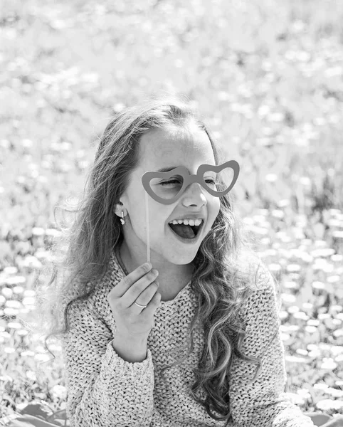 Amor desde el concepto de primera vista. Niño posando con gafas en forma de corazón de cartón. Chica se sienta en la hierba en Grassplot, fondo verde. Chica en cara feliz pasar el ocio al aire libre —  Fotos de Stock