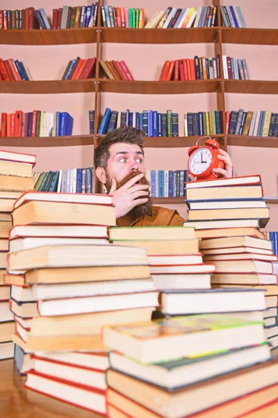 Άνθρωπος, επιστήμονας που κρυφοκοιτάζει από σωρούς από βιβλία με ξυπνητήρι. Δάσκαλος ή μαθητής με τη γενειάδα μελέτη στη βιβλιοθήκη. Προθεσμία έννοια. Ο άνθρωπος στο πρόσωπο έκπληξη που βλέπουν ρολόι, ράφια σε φόντο — Φωτογραφία Αρχείου