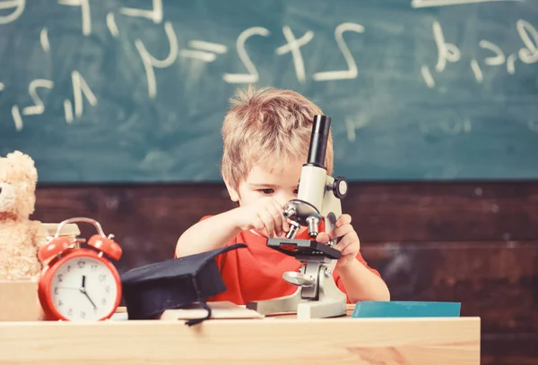 Junge arbeiten mit Mikroskop im Klassenzimmer, Kreidetafel im Hintergrund. Kind auf beschäftigtem Gesicht nahe Uhr und Teddybär. Grundschulkonzept. Erste Ehemalige interessiert an Studium, Lernen, Bildung — Stockfoto