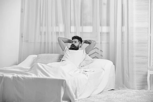 Macho con barba cogido de la mano detrás de la cabeza, relajante. Hombre de camisa acostado en la cama, cortinas blancas en el fondo. Un tipo bostezando la cara lleno de energía en la mañana. Lleno de fuerza y concepto de energía —  Fotos de Stock