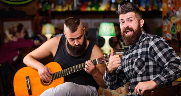 Hipster brutal bearded with friend in pub. Man play guitar in pub. Cheerful friends sing song guitar music. Relaxation in pub. Friends relaxing in pub. Live music concert. Acoustic performance in pub