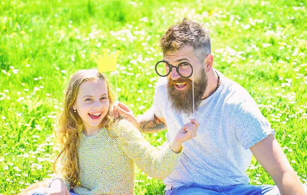 O pai e a filha sentam-se na relva no campo, fundo verde. Conceito de princesa Daddys. Criança e pai posando com coroa e óculos atributos de cabine de fotos. Família passar o lazer ao ar livre — Fotografia de Stock