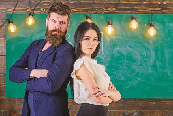 Man with beard and teacher in eyeglasses stand back to back, chalkboard on background. Teacher and schoolmaster look confident. Lady and hipster working together in school. School staff concept