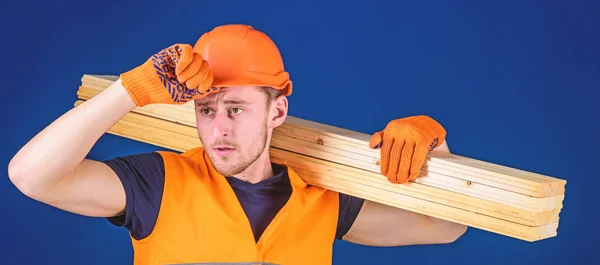 Construction worker carrying a wood plank on his shoulder. Carpenter wearing hard hat, protective gloves and safety orange vest, blue background. Protective equipment concept — Stock Photo, Image