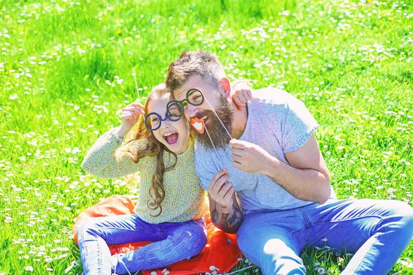 Niño y padre posando con anteojos atributos fotomatón en el prado. Concepto inteligente e inteligente. Papá y su hija se sientan en la hierba en Grassplot, fondo verde. Familia pasar el ocio al aire libre — Foto de Stock